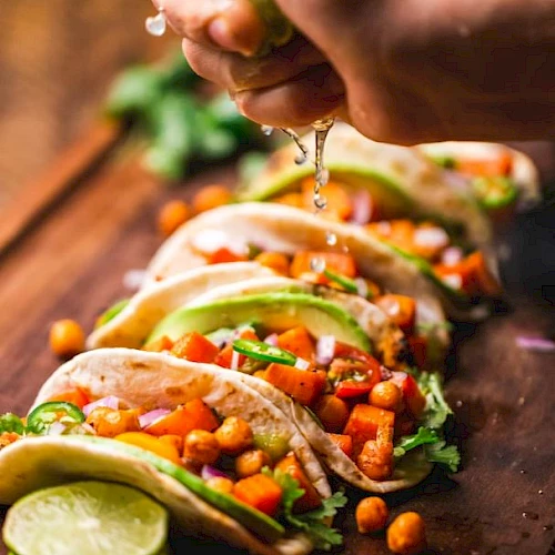 A hand squeezes lime over tacos filled with vegetables and garnished with avocado, on a wooden board with lime slices nearby.