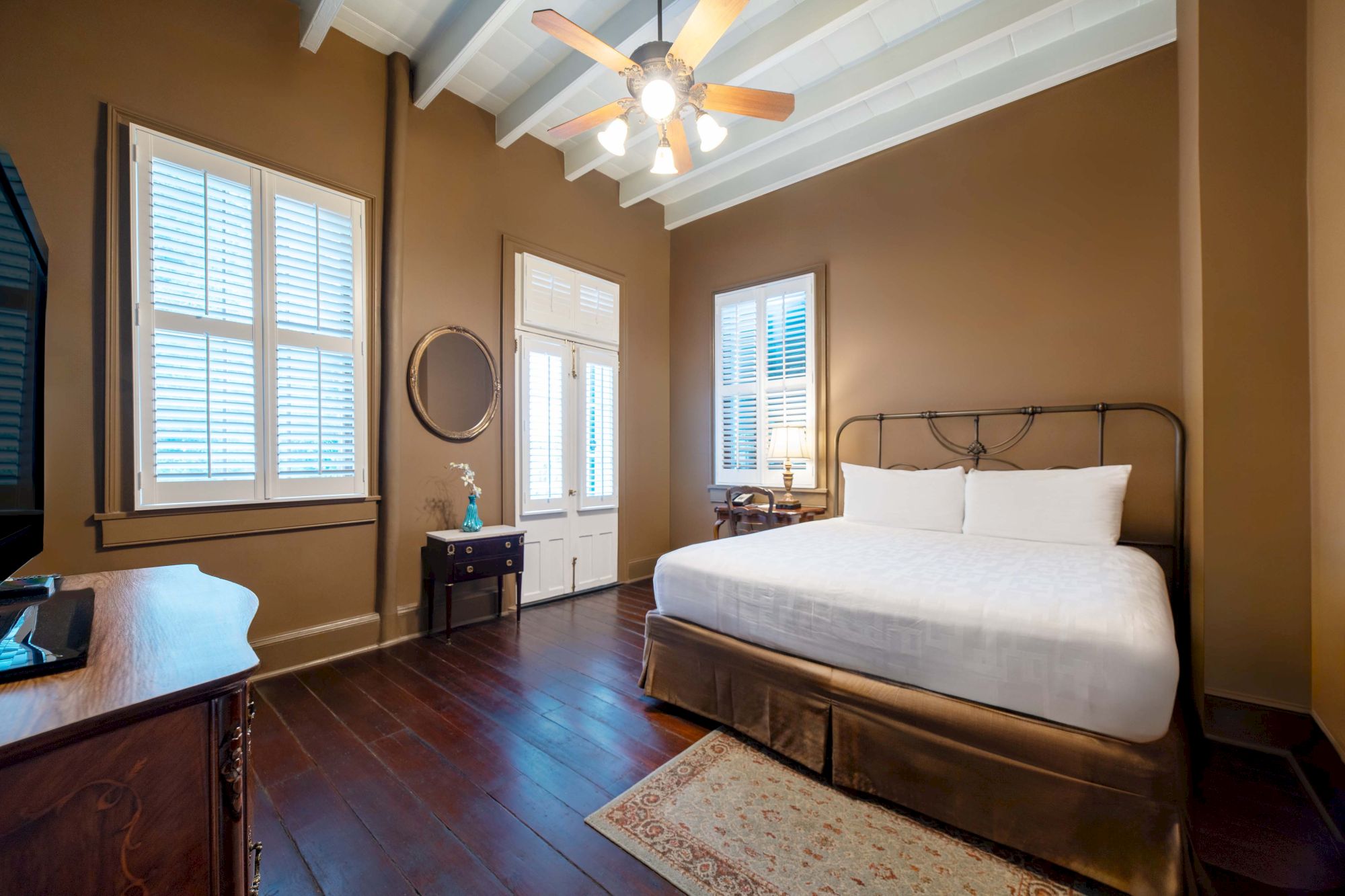 A cozy bedroom with a large bed, wooden floors, a ceiling fan, and natural light from two windows with shutters, creating a warm ambiance.