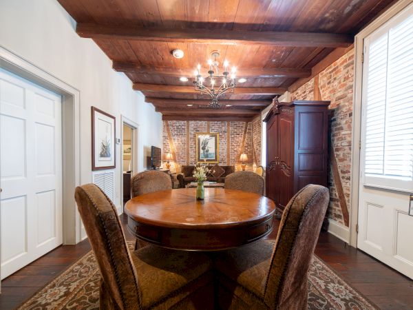 A cozy dining room with a round wooden table, plush chairs, brick walls, wooden ceiling beams, and a chandelier above the table.