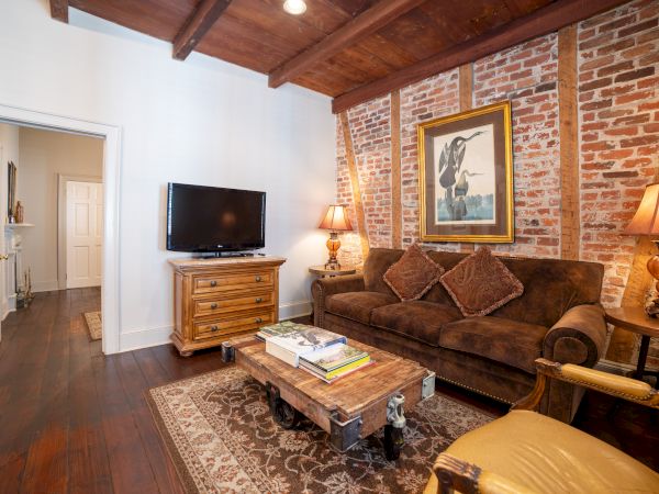 A cozy room with a brown sofa, exposed brick wall, artwork, TV, wooden beams, lamps, and a coffee table on a rug.