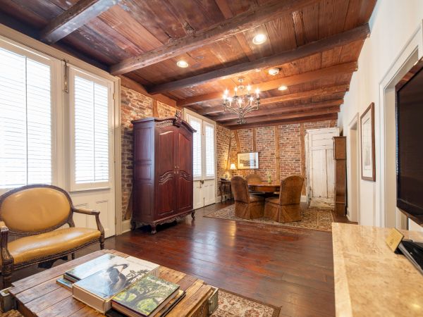 A rustic living room features wooden beams, brick walls, a chandelier, vintage furniture, and a TV. Books are on a wooden coffee table.