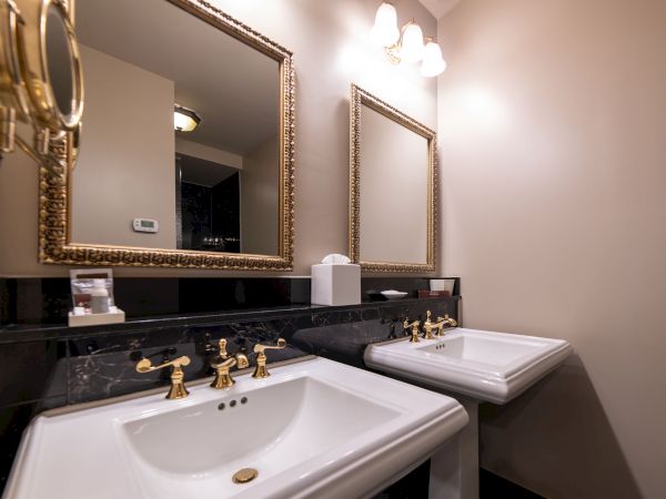The image shows a bathroom with two sinks, ornate mirrors, gold fixtures, and a towel dispenser on a black counter.