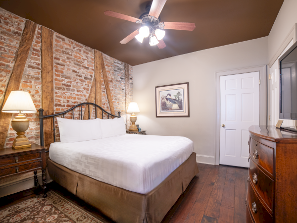 A cozy bedroom with a brick accent wall, double bed, bedside tables with lamps, framed art, wooden dresser, ceiling fan, and TV.