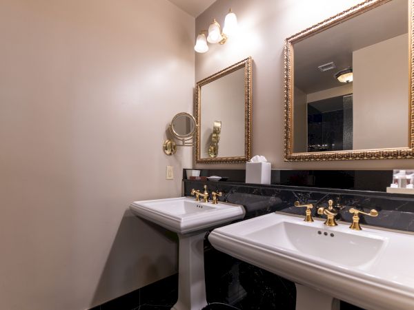 The image shows a bathroom with two pedestal sinks, gold fixtures, two framed mirrors, and wall lamps above.