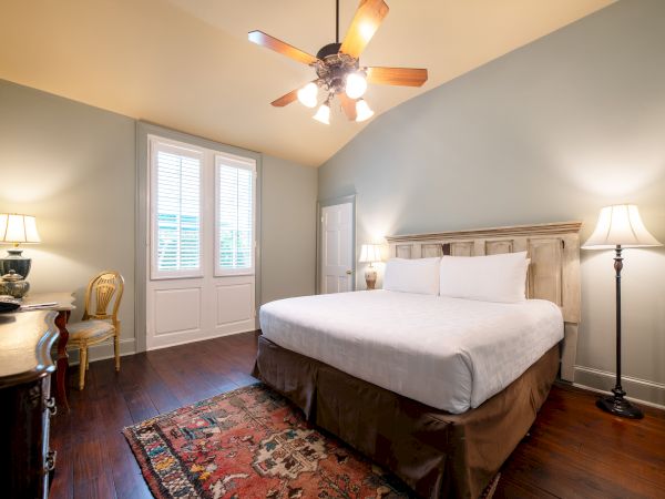 The image shows a bedroom with a large bed, wooden furniture, a ceiling fan, lamps, and a patterned rug on a wooden floor.