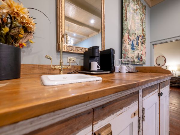 A rustic kitchen counter with a sink, coffee maker, cups, and a vase of flowers, near a mirror and a decorative wall hanging.