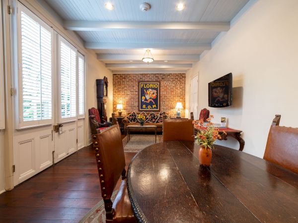 The image shows a cozy living room with a wooden table, leather chairs, floral decorations, a brick accent wall, and wall-mounted art.