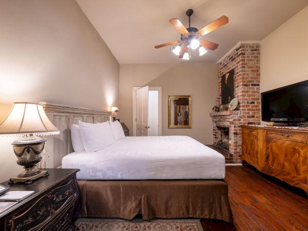 A cozy bedroom with a bed, vintage furniture, a brick fireplace, lamp, ceiling fan, and a TV on a wooden dresser, creating a warm ambiance.
