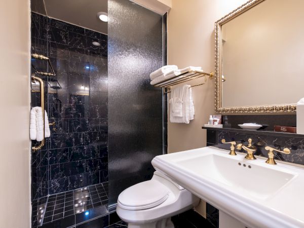 The image shows a bathroom with a black tiled shower, a toilet, a sink with gold fixtures, a large mirror, and a shelf with towels and toiletries.
