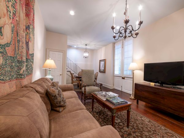 A cozy living room with a brown sofa, tapestry, chandelier, TV, and armchairs. Stairs lead to the next floor, and light filters in.