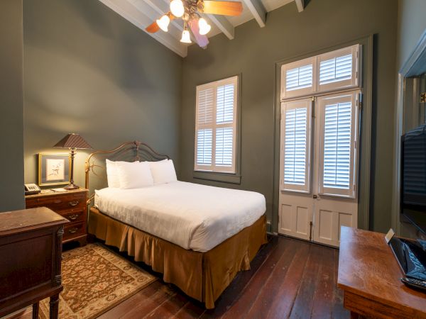 A cozy bedroom with a double bed, wooden furniture, a ceiling fan, and natural light coming through the window shutters.