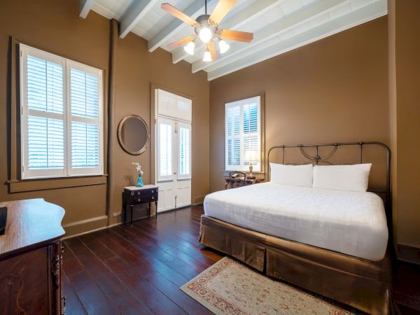 A cozy bedroom with a large bed, brown walls, wooden floors, ceiling fan, and windows with shutters. A dresser and mirror are also present.