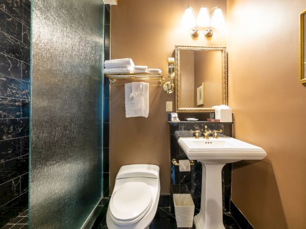 A bathroom featuring a toilet, pedestal sink, mirror, towel shelf, and glass shower, warmly lit with wall-mounted lights.