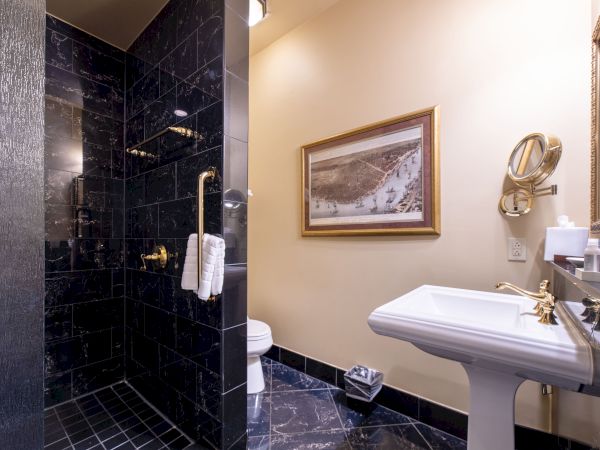 The image shows a bathroom with black tiled walls, a shower area, a pedestal sink, a toilet, and a framed picture on a beige wall.