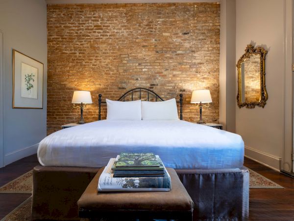 A cozy bedroom features a bed with white linens, exposed brick wall, two lamps, a decorative mirror, and a stack of books on a bench.