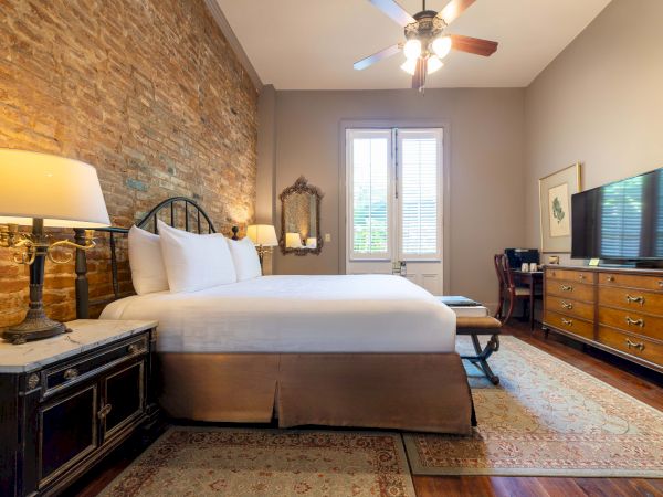 A cozy bedroom with a bed, brick accent wall, two lamps, TV on a dresser, and ceiling fan, featuring natural light from large windows.