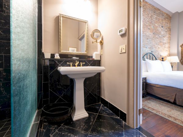 A bathroom with a pedestal sink and mirror, next to a bedroom with a brick wall and bed.