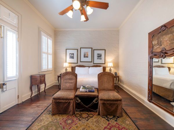 A cozy bedroom with a bed, framed pictures, chairs, a rug, and a large ornate mirror, illuminated by a ceiling light fixture.