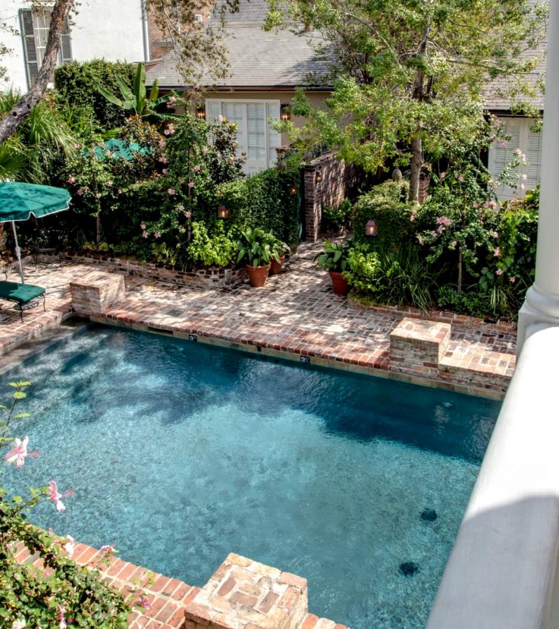 A swimming pool with clear blue water surrounded by brick pavers, lounge chairs, and lush greenery.