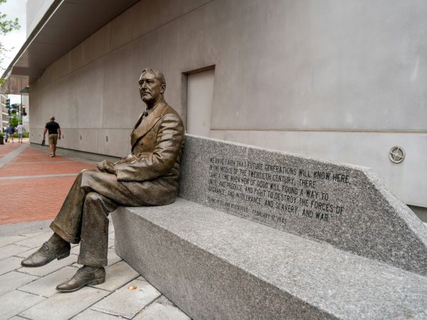 A statue of a seated man is integrated into a stone bench with engraved text, located on a city sidewalk.