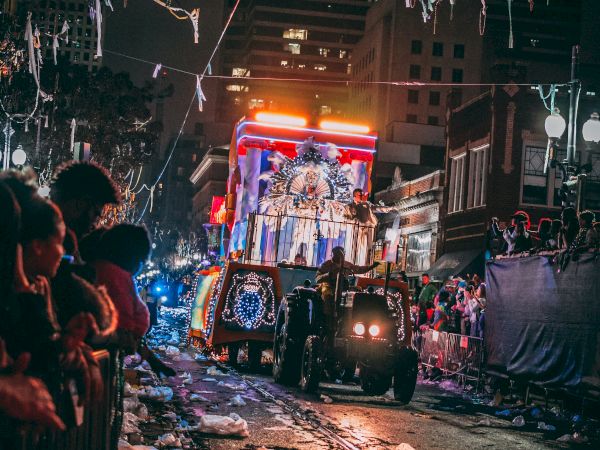 A nighttime parade with a decorated float passes through a crowd-lined street, colorful lights illuminating the scene.