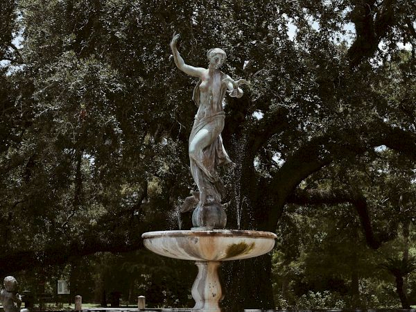 A statue of a person stands atop a multi-tiered fountain surrounded by trees and greenery in a park setting.