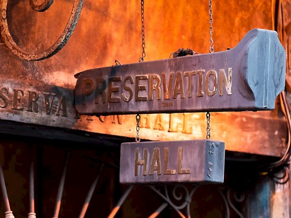 The image shows a vintage sign for "Preservation Hall," hanging from a building with rustic metalwork and an aged appearance.