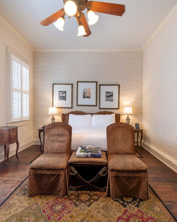 A cozy bedroom with a bed, two chairs, a wooden floor, a large mirror, and framed art on the wall, in warm lighting.