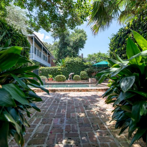 A brick path leads to a pool, surrounded by lush greenery and a house in the background under a clear blue sky.