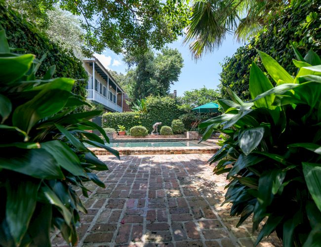 A brick path leads to a pool, surrounded by lush greenery and a house in the background under a clear blue sky.