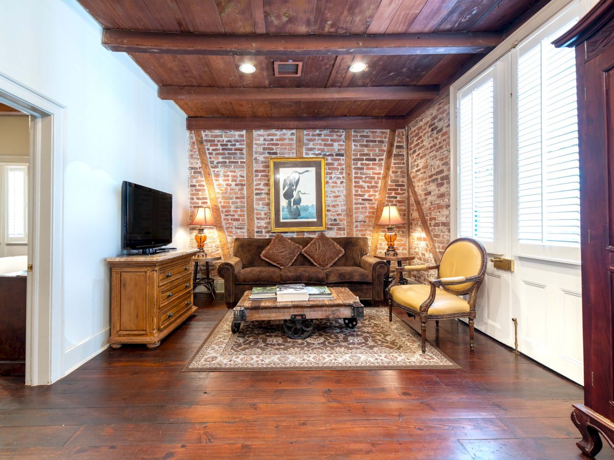 A cozy living room with brick walls, wooden ceiling, sofa, chairs, TV, and art. Adjacent to a bedroom with a door.