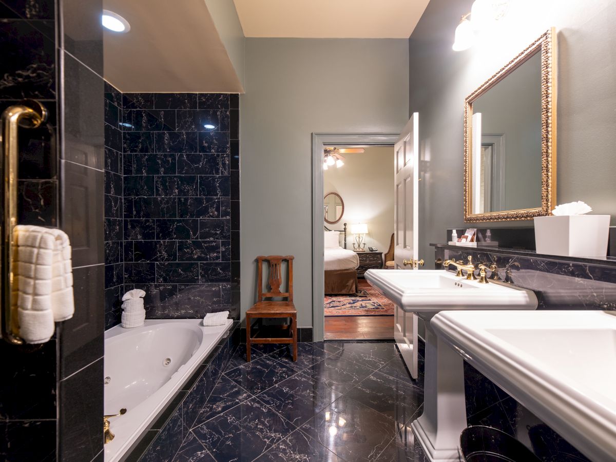 A luxurious bathroom features black marble tiles, a bathtub, dual sinks with mirrors, and a glimpse of a bedroom through an open door.