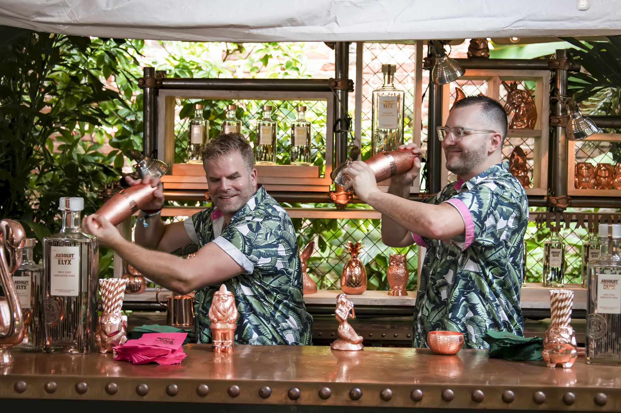 Two bartenders in hawaiian shirts mix cocktails behind a copper bar, juggling shakers, with copper sculptures on the shelf behind them.