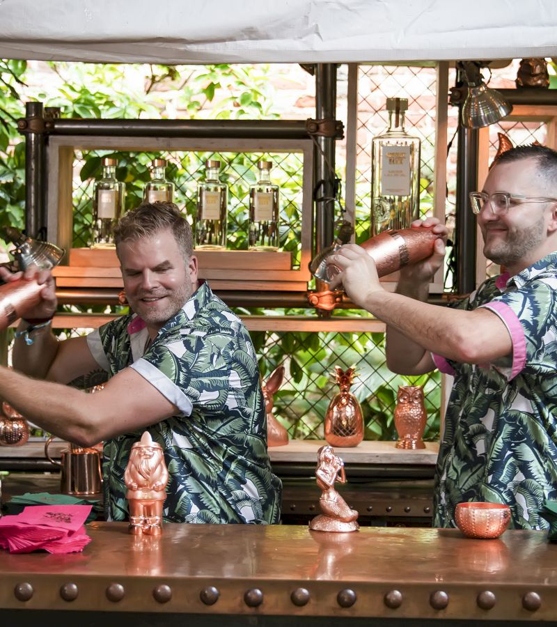 Two bartenders in Hawaiian shirts shaking cocktails at a copper bar with tropical decor.