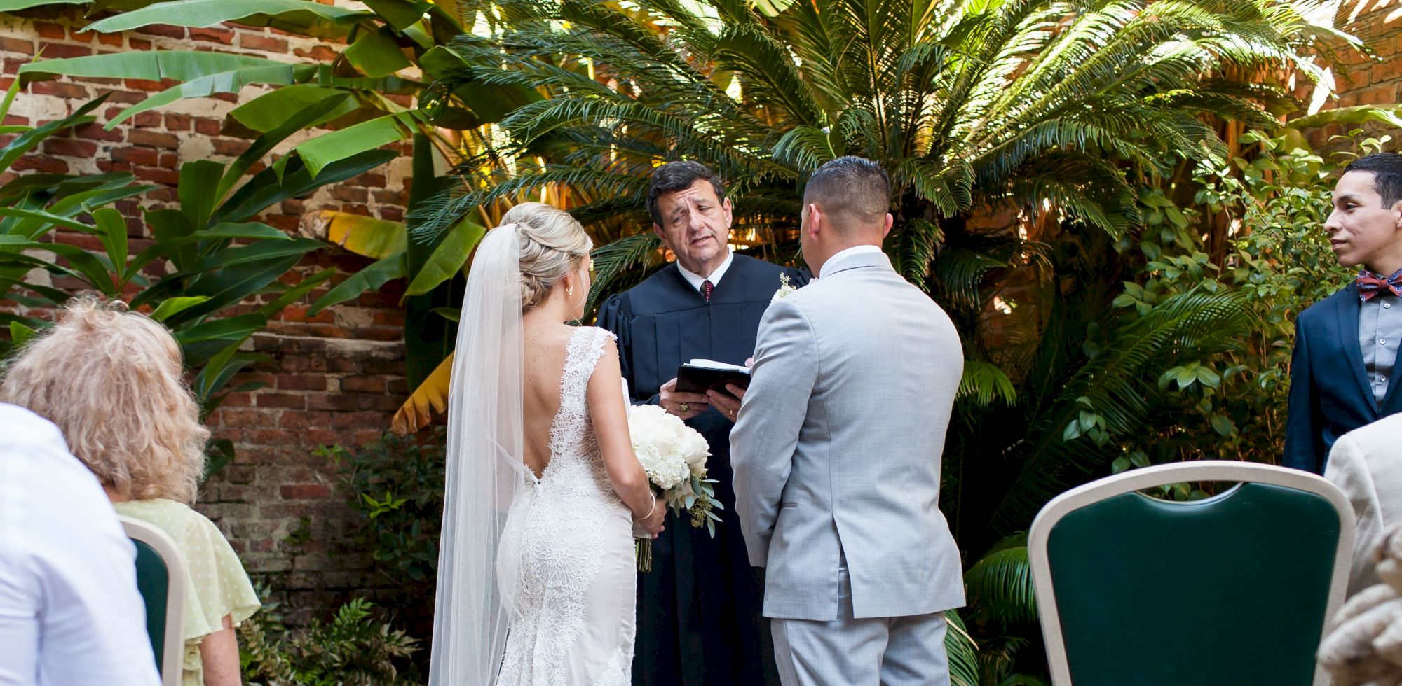 A couple is getting married outdoors with a small audience, surrounded by lush greenery and brick walls.