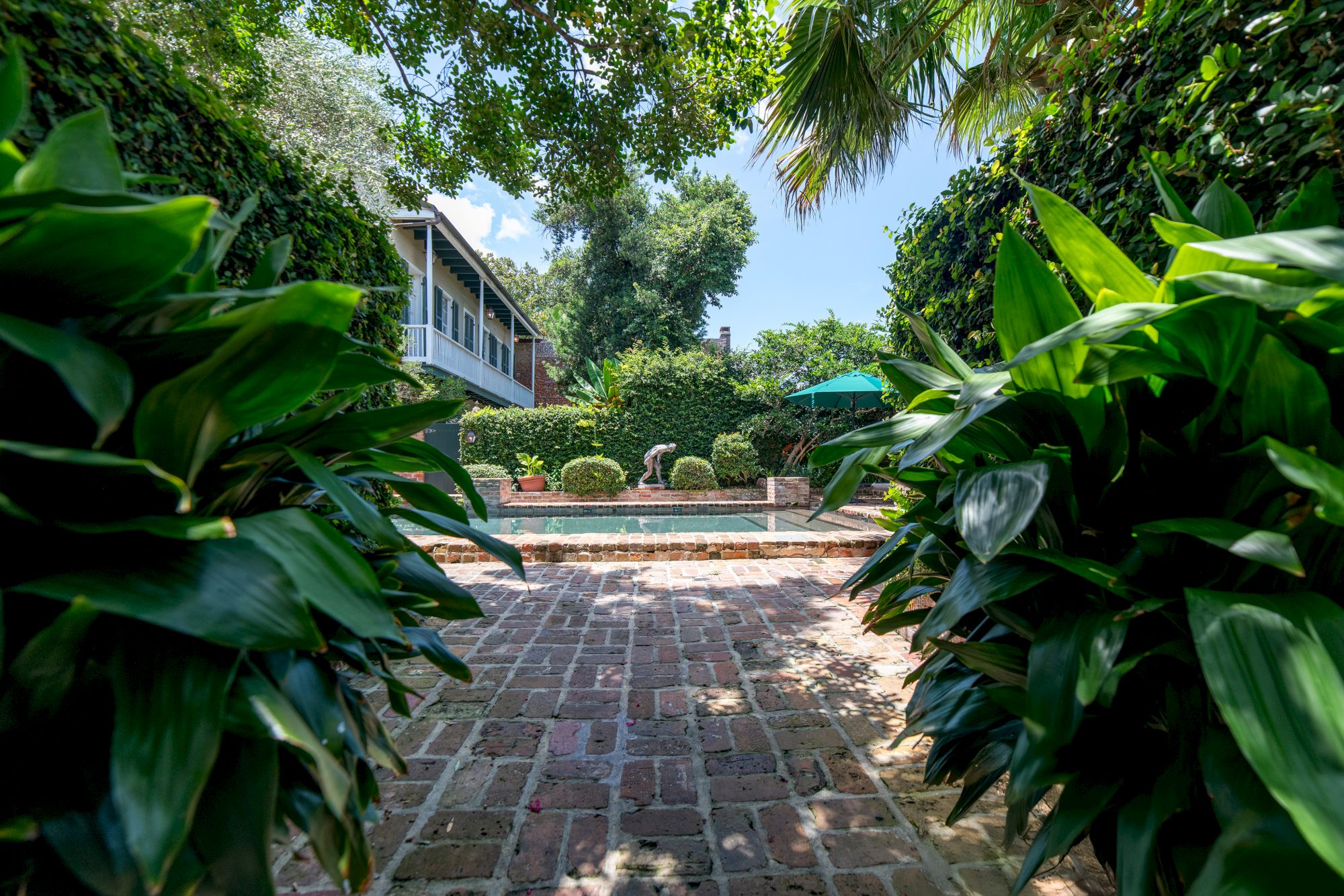 Lush garden path with brick paving leads to a seating area and greenery, surrounded by leafy plants and trees, with a building nearby.