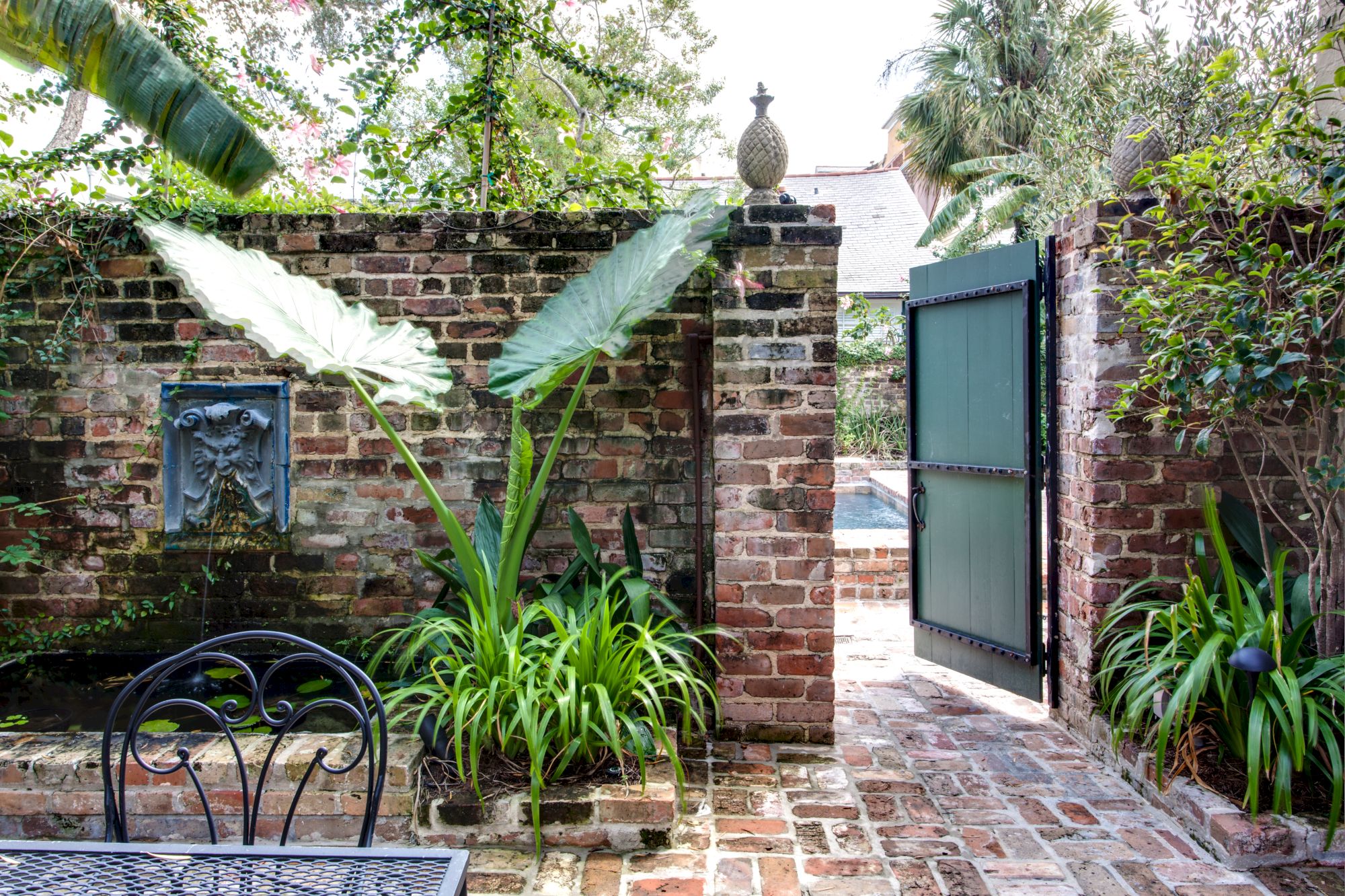 A brick courtyard features lush greenery, a wrought iron table, and an open gate leading to a pool area, creating a serene escape.
