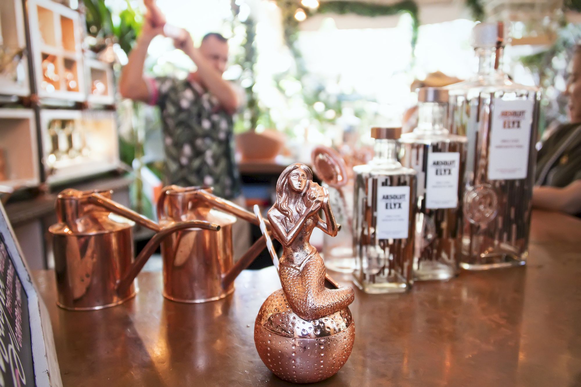 A copper bar setup with a small copper teapot in the foreground, bottle jars labeled &ldquo;AMU,&rdquo; mugs, and a blurred bartender behind the counter.
