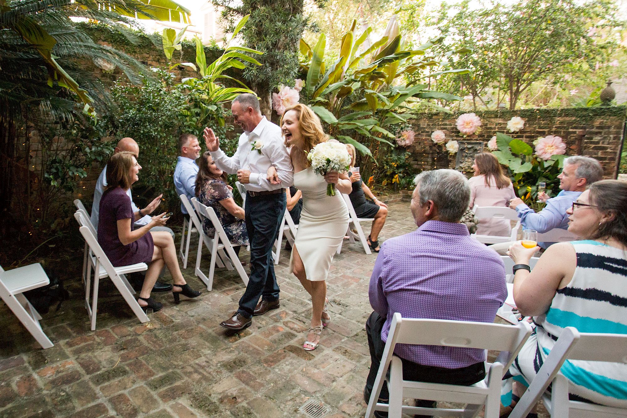 A couple joyfully walks down an aisle outdoors, surrounded by seated guests on white chairs, in a garden setting.