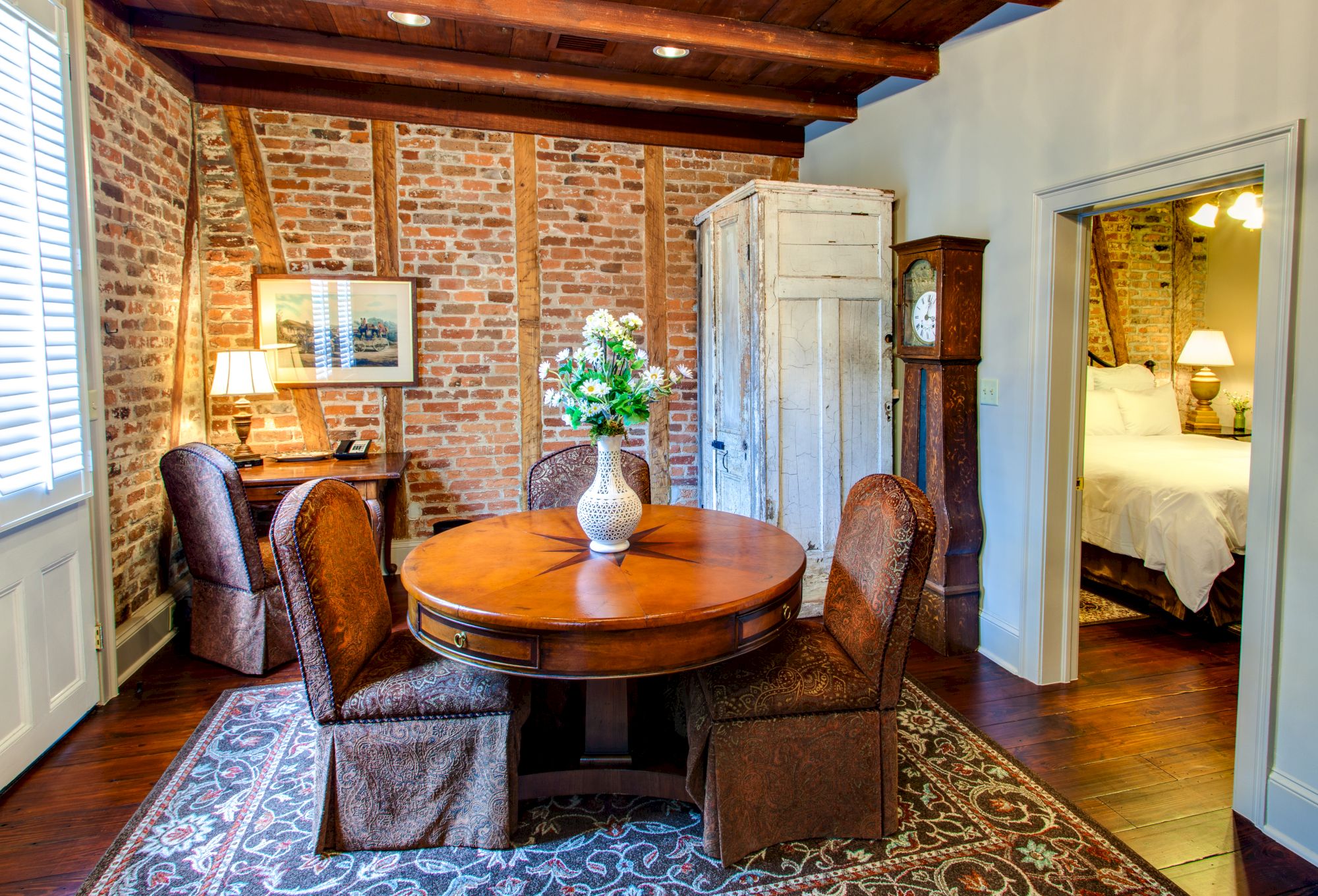 A cozy room features a round wooden table, brick walls, a vase with flowers, and a glimpse of a bedroom through an open door.