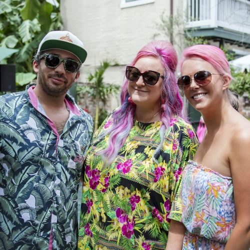 Three friends pose by a poolside in tropical outfits; two women with pink hair and sunglasses stand with a man in a patterned shirt and cap.