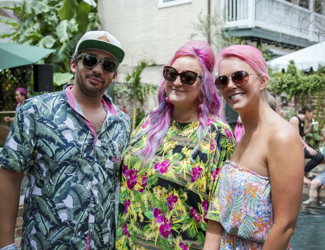 Three friends pose by a poolside in tropical outfits; two women with pink hair and sunglasses stand with a man in a patterned shirt and cap.