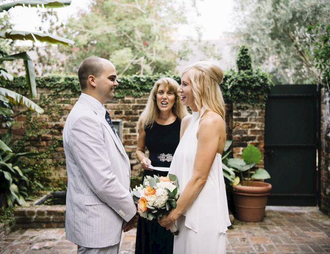 A couple is standing together with a woman officiating, in a garden setting. The scene appears to be a wedding ceremony.