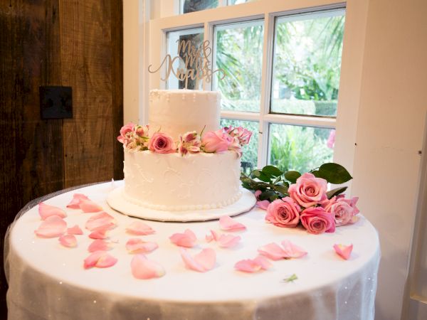 A two-tier white cake with pink roses and petals on a table, set near a window, creating a romantic ambiance.