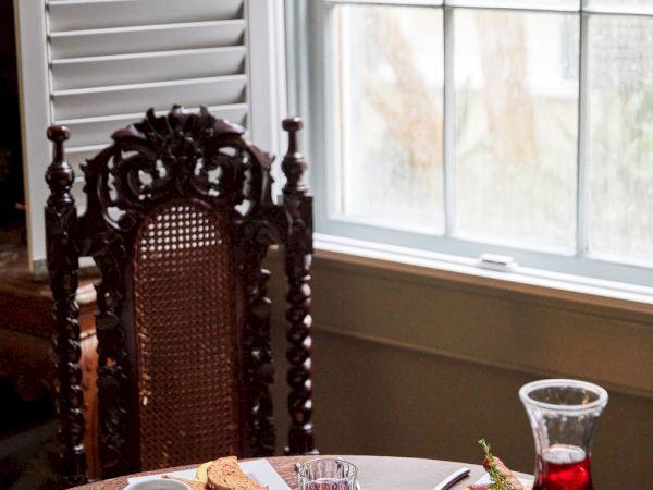 A cozy breakfast scene with a slice of toast, fruit, a newspaper, and a carafe of red drink by a window, next to an ornate chair.
