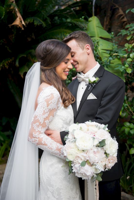 A bride and groom are embracing in a garden setting, the bride holding a bouquet of flowers, both wearing formal wedding attire.