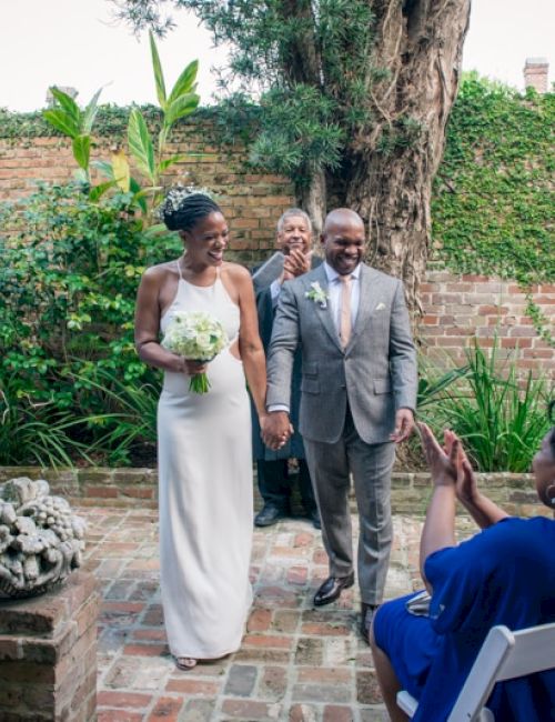 A couple is walking hand in hand at an outdoor wedding ceremony, with guests seated and clapping in a garden setting.