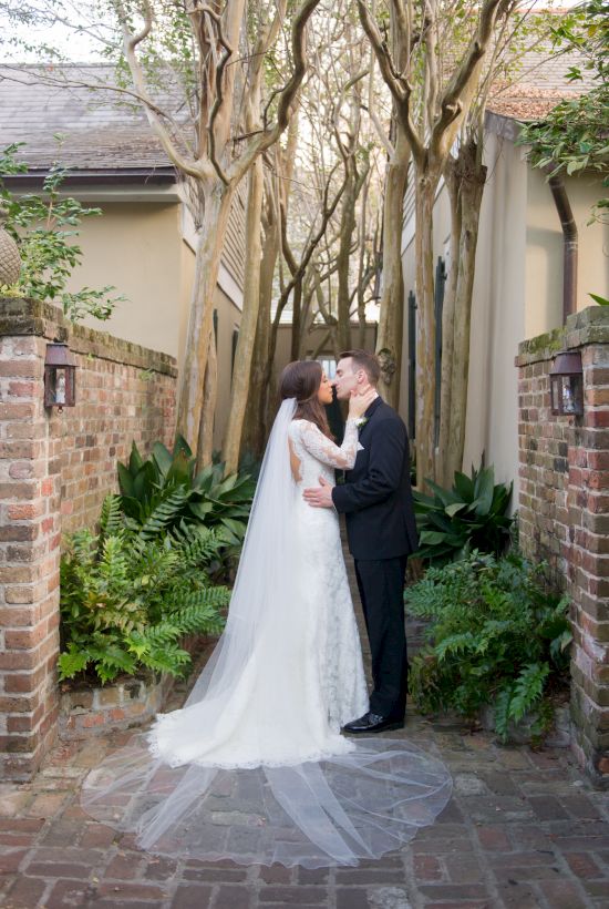 A bride and groom are kissing in a garden pathway, surrounded by trees and brick walls, with the bride in a white dress.