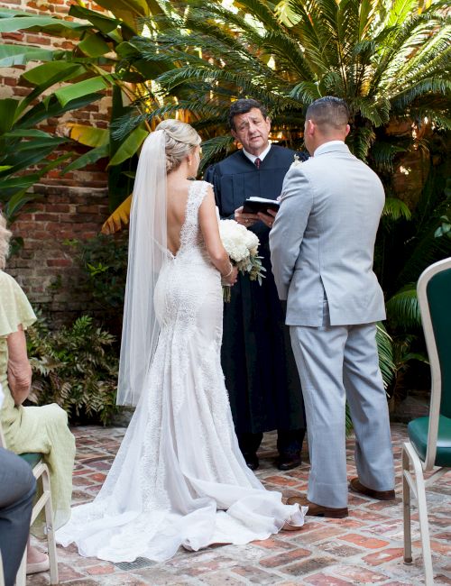 A couple stands at an outdoor wedding ceremony with guests seated. The setting includes greenery and brick walls, creating a cozy ambiance.