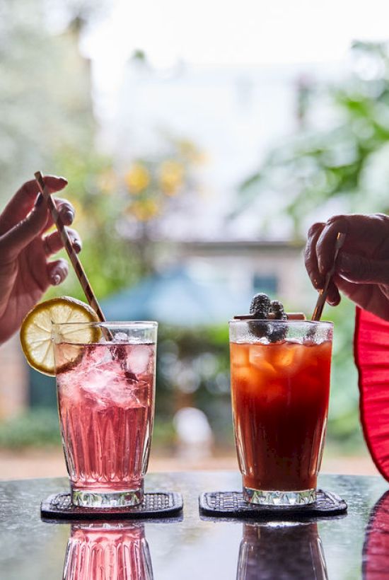 Two people are toasting with colorful cocktails, garnished with citrus and berries, outdoors on a reflective table.